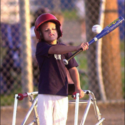A young boy stands on a baseball field. He is wearing a red helmet, a black shirt, and white baseball pants. He is using a walker mobility device. He is swinging a blue baseball bat at a white baseball.