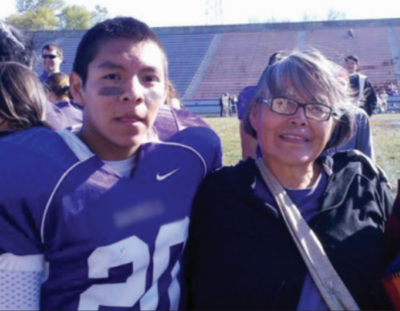 A son and mother stand together on the side of a football field, smiling at the camera. They are both Native American. The son is on the left in a purple football jersey with lines painted under his eyes. The mother is on the right with short gray hair and glasses. She is wearing a purple shirt and a black jacket.