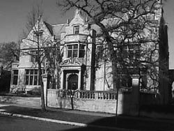 Grayscale image of a large, historic-looking building. It is at least two stories high and has a short, stone fence. The building is made of stone and has many windows. 