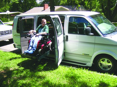A teenage boy in a power chair on a wheelchair lift into a van smiles at the camera. He is wearing a green t-shirt and jeans. The van is parked on a curb next to a green lawn.