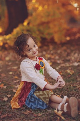 A young female child in a white shirt and multi-colored shirt sits on the grass with her arms in front of her and her legs crossed. She has short brown hair and she is smiling. Behind her is a tree along with many red, yellow, and green leaves. 