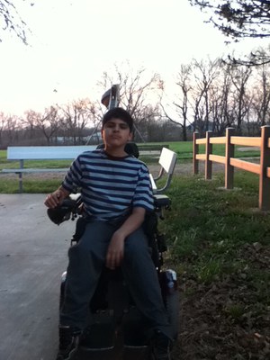 A young man with dark hair in a power chair is in front of grass and a wooden fence. He is wearing a striped shirt and jeans and is looking at the camera.