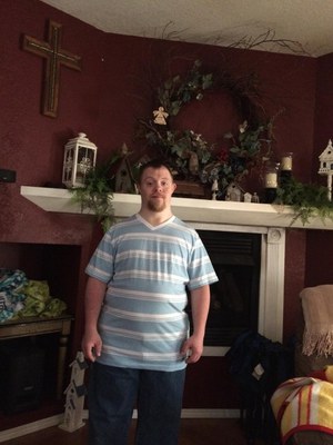 A man with Down Syndrome stands in front of a fireplace. There is a wreath, a cross, and other decorations on the mantle. He has brown hair and a beard and is wearing a striped blue and white shirt and jeans.