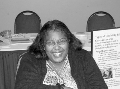 Black and white photo of a woman sitting indoors. She is smiling at the camera. She has dark hair and skin and thin wire glasses. She is wearing a button down shirt and a cardigan.
