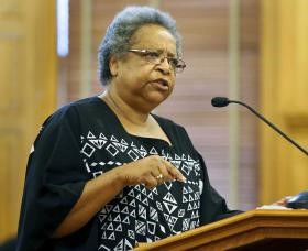 A Black woman with gray hair and glasses is speaking at a podium into a microphone. She is wearing a black and white dress. She is pointing one finger at the podium and she has an expression of passion.