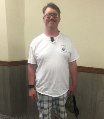 A middle-aged white man with a mustache and glasses stands in front of a wall. He is wearing a white shirt, plaid shorts and a watch. He has a hat in his hand. He is smiling at the camera.