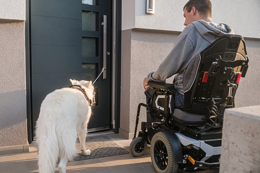Stock image of a man using an electric wheelchair with his service animal entering an apartment building.