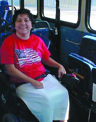 A woman is sitting on a bus seat. She is wearing her seatbelt. She has short brown hair and is smiling at the camera. She is wearing a red shirt with an American flag on it and a long white skirt. 