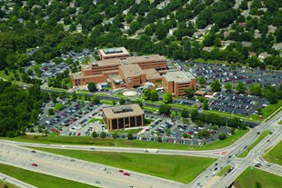Birdseye view of a hospital facility. It is surrounded immediately by parking lot and then by trees.