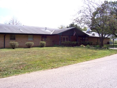 Image of a brick facility building and grass lawn. Three shrubs are in front of the building. The building has a door and windows and appears as one story.