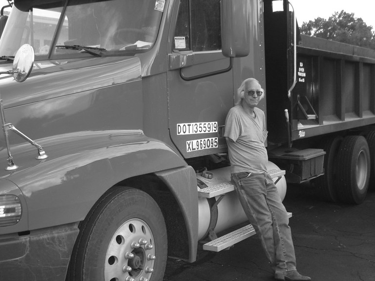 Grayscale image of a man leaning against a large truck. He is wearing a t-shirt and jeans. He does not have a right arm. He has shoulder-length grey hair and is wearing dark sunglasses.