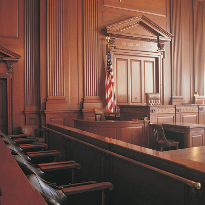 Image of a courtroom. The walls have wood paneling and behind the judges chair states “In God We Trust.” The witness stand on the left is visible and has an American flag behind it. One row of seats is shown in the image. 