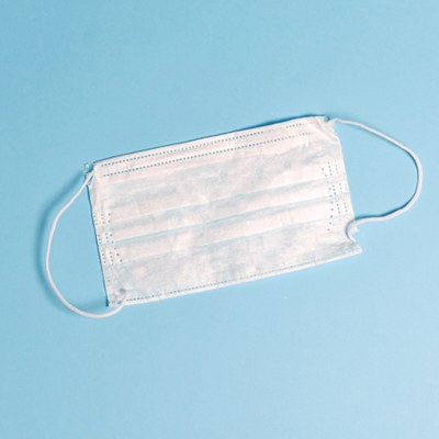 This is an image with a plain, sky blue background. On top of the background is one disposable white face mask that is laid out flat.  