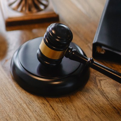 This is an image of a wood gavel. The gavel is resting on a circular, raised piece of dark wood. It is on top of a table. The gavel itself has a gold strip around the middle. Part of a black binder is shown next to the gavel. 
