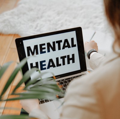 This is an image from behind a woman’s shoulder showing only her laptop screen, which states “MENTAL HEALTH.” She has brown hair and only part of her beige blazer is visible, along with her black watch. 