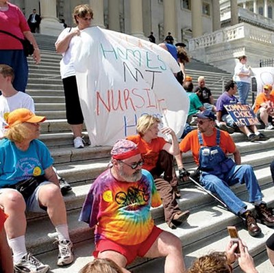 Image of a disability rights protest at the U.S. capitol building. Around 15 people are sitting on the Capitol steps. Two people are holding up a sheet that has something unreadable written on it.  Other people are holding posters in the background. 