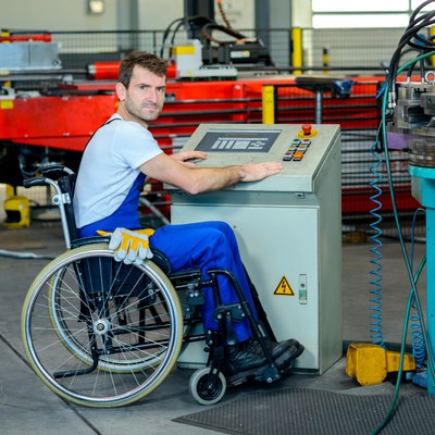 A white adult man using a wheelchair is sitting in front of an operating board in a mechanical shop. He is wearing blue overalls and a white t-shirt. He has gloves sitting on his lap.