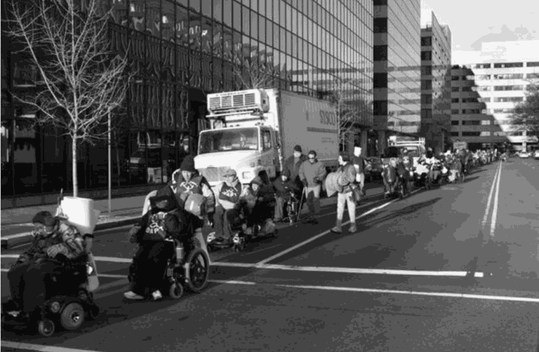 A line of people move through a city street with tall buildings on the side. Two people using power chairs lead the march. Behind them are many more people - some walking, some using power chairs, and some using manual wheelchairs.