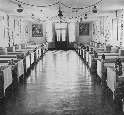 Black and white image of a large room with beds lining both walls. The room is decorated with posters. A stuffed animal is on each bed.
