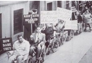 This is a black and white photograph of about twenty disabled veterans in wheelchairs. The men are waiting in a line on the sidewalk along the wall of an office building. The wall looks like the façade of a government building, with smooth grey stone and tall, barred windows. The men wear clothes of the 1940s and 1950s: wide lapels, white socks and dark shoes, cardigan sweaters. One man smokes a pipe and is wearing a suit with a wide striped tie and Argyll socks. The men hold signs that say: "How Dare They?"; "Keep Faith in Disabled Vets"; "How About the East Orange VA Hospital"; "We Did Our Share, Treat Us Right": "PVA"; "Don't Break Promises".