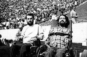 Black and white image of two men in wheelchairs. Both men have dark hair with beards and mustaches. There is a crowd behind them. 
