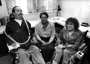 Black and white image of three people in an indoor room. On the left is Ed Roberts. He is using a wheelchair and a breathing device that goes from his mouth to behind his back. He has dark hair and a mustache. He is wearing khaki pants and a dark button up shirt. In the middle is a woman with her hair pulled back. She is wearing a button down shirt and a long skirt. On the right is Judy Huemann. She is wearing a button down shirt and dress pants. She has short, curly hair and is using a wheelchair.