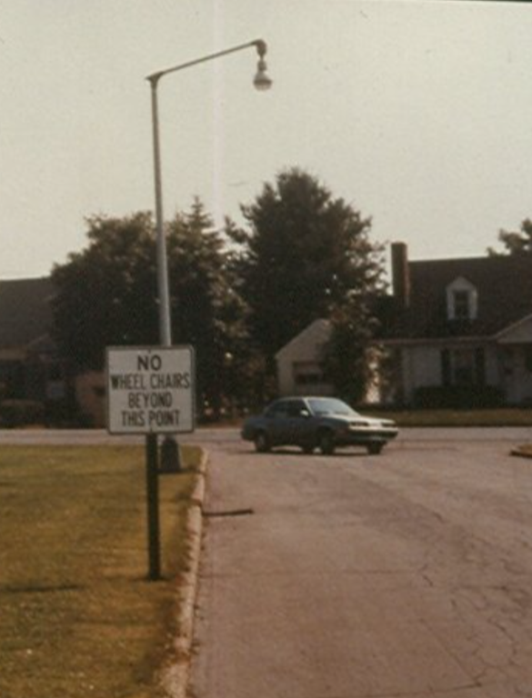 This is a faded color photograph showing a flat suburban neighborhood street. It is quiet and deserted. There is no sidewalk but a slightly worn grassy footpath edges the side of the street. A flat area of close cut grass extends out from the curb along the street. There is a street light on a high pole. In the background rises a two-story brick building and two white houses with big trees in the front yards. TV aerials are visible on rooftops in the distance. A car is just rounding the corner at the far end of the street and headed this way, perhaps into a parking lot or down another street. There is a white metal sign with black letters placed at the curb that reads: "No Wheel Chairs Beyond this Point."