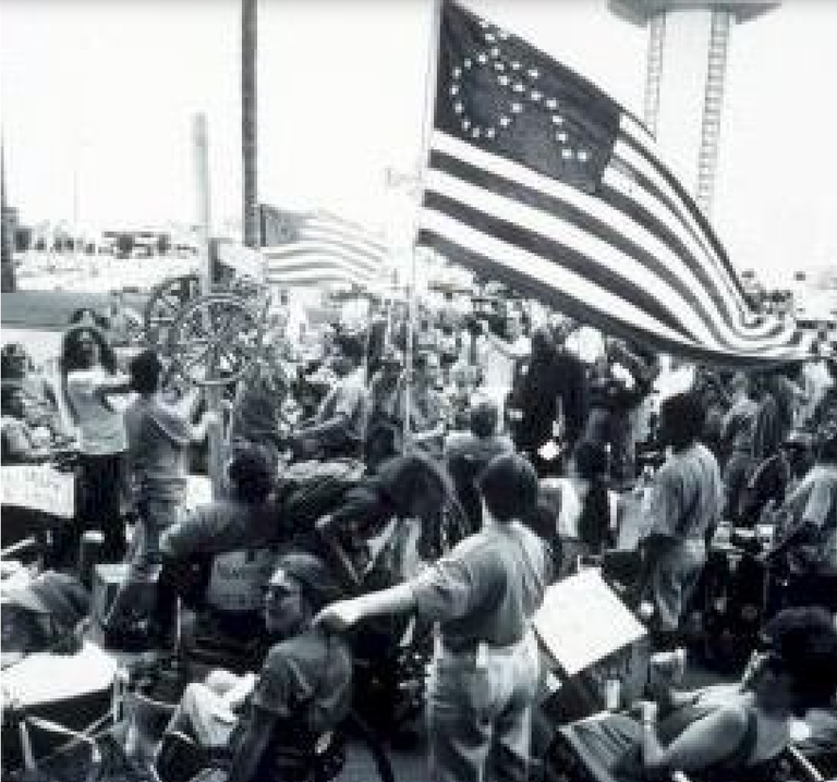 Black and white image of a crowd of people. A large flag is waving. It is designed similarly to the U.S. American flag, with stripes and a navy blue rectangle in the upper right corner. Instead of white stars in rows, stars form the shape of a person in a wheelchair.