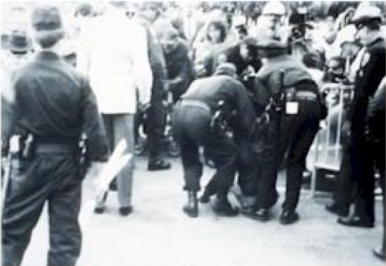Black and white photo. Two police officers are arresting someone who is not visible in the image. They are leaning toward the ground.