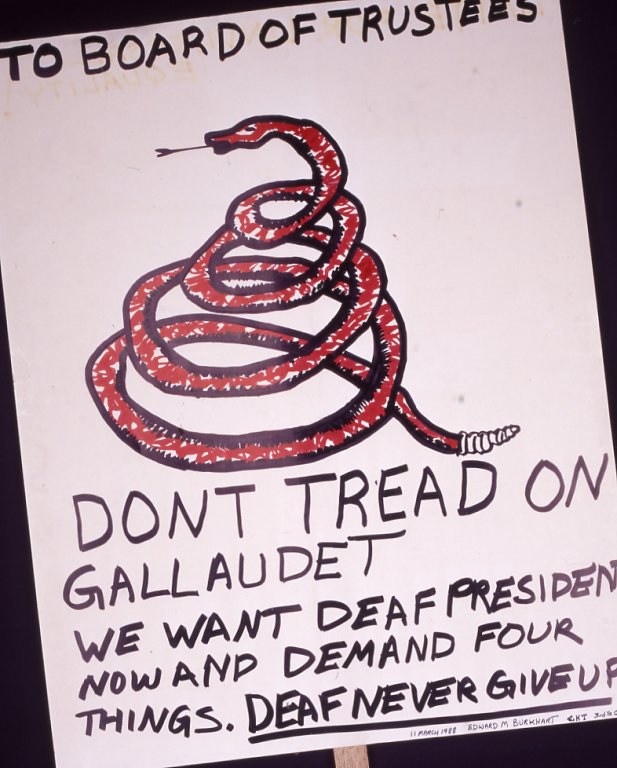 A white poster is on a wooden stick. At the top it says "TO BOARD OF TRUSTEES." Below that text is a drawing of a red, coiled snake. Underneath the text, handwriting reads: "DON'T TREAD ON GALLAUDET. WE WANT DEAF PRESIDENT NOW AND DEMAND FOUR THINGS. DEAF NEVER GIVE UP"
