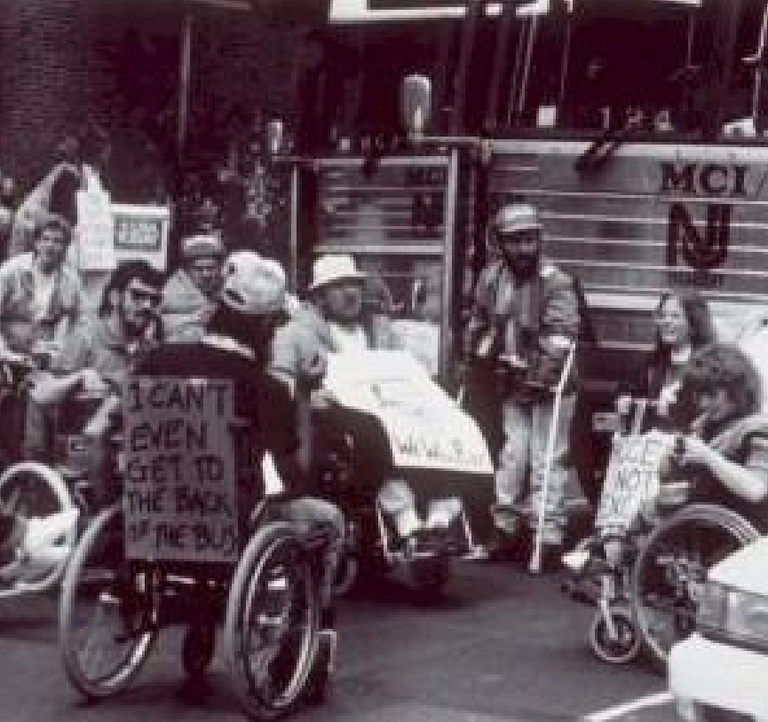 Black and white image of people in wheelchairs and other mobility assisting devices in a parking lot. Someone is in a wheelchair closest to the camera with a sign on the back of the chair that reads "I CAN'T EVEN GET TO THE BACK OF THE BUS." Other people have signs that are unreadable in the image.