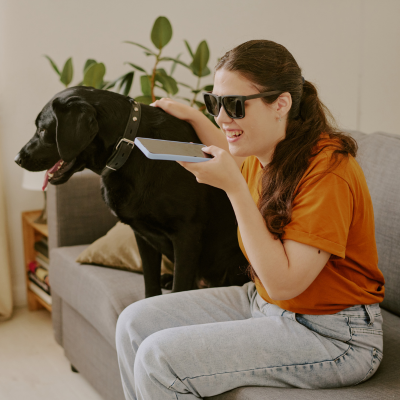 Stock photo of young woman with a visual impairment on the phone in her apartment, sitting next to her service dog on the couch. 