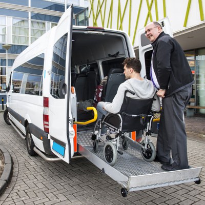 A white adult man is pushing another white adult man’s wheelchair up a metal ramp into a white van. The van is outside of a building on a driveway path.