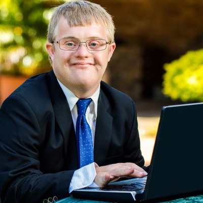 A white adult man with a developmental disability smiles at the camera. He is blonde and is wearing glasses. He is also wearing a suit jacket, white button up shirt, and a blue tie. He is using a black laptop on a table. 