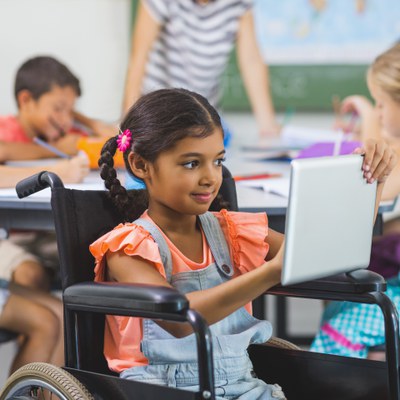 An elementary school-age, Indian, female student with braids and a flower clip in her hair is sitting in a wheelchair and using a tablet device. She is holding up the tablet and smiling. She is wearing an orange shirt and light blue overalls.