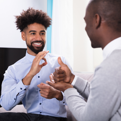 Photo of two Black men facing each other in sitting position. The man on the right has a beard and mustache and is wearing a blue button-up shirt and black slacks. He is signing in ASL toward the other man. The second man’s face is not visible to the camera but he is wearing a white collared button down with a grey sweater. He is also signing in ASL toward the other man.