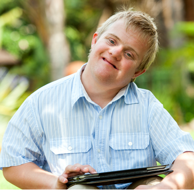 A white adult man with a developmental disability smiles at the camera. He is blonde and is wearing a light blue, button-up, short sleeve shirt with white pinstripes. He is holding a tablet and is touching the screen with his right index finger. The background is trees and plants but is blurred. 