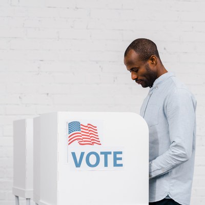 A Black adult man is standing and looking down over a polling station. The polling station has a sticker that says “VOTE” with an American flag. The man has short hair and a stubble. He is wearing a button down, light blue shirt. 